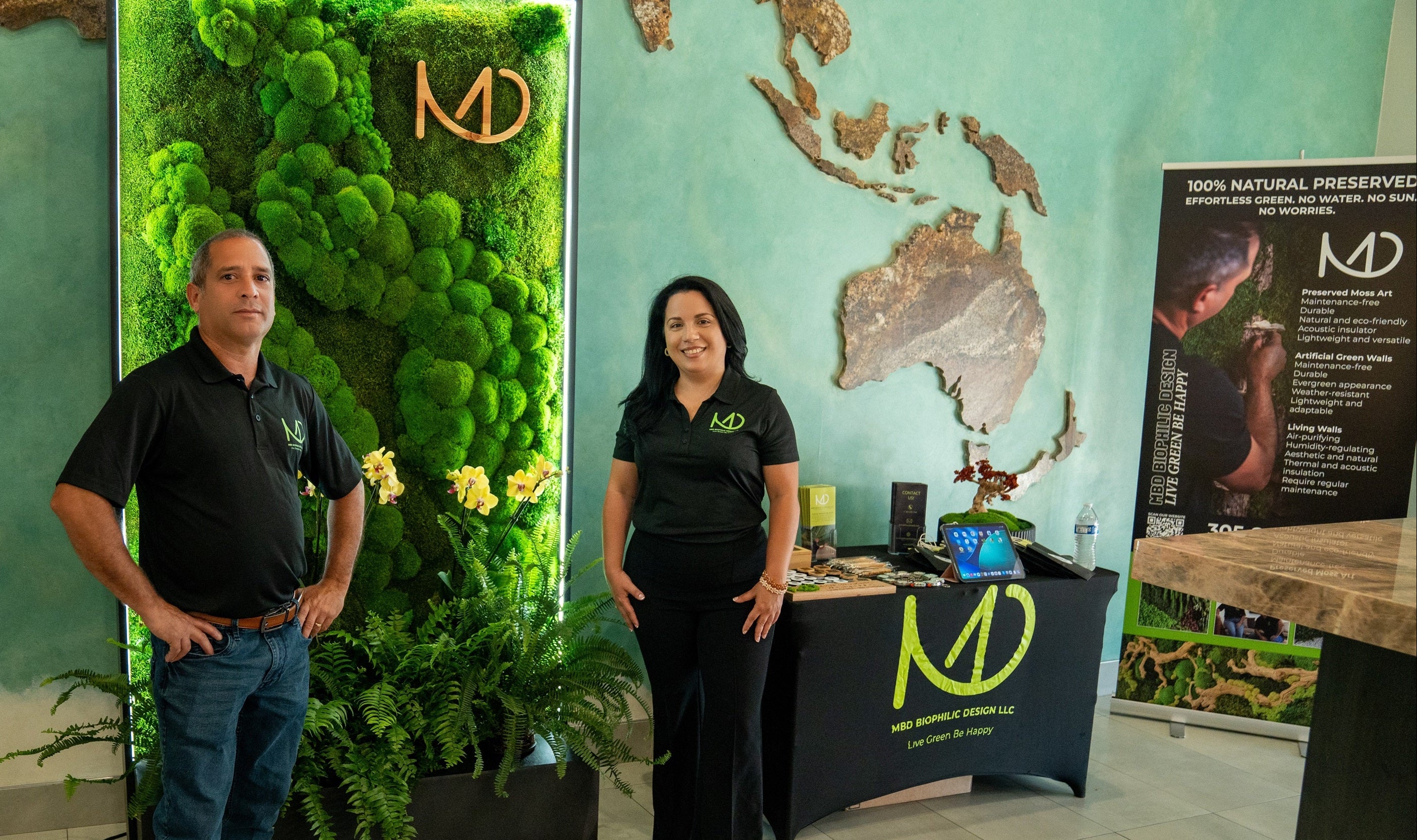 Two people standing in front of a green wall with a logo and a map on a teal wall.